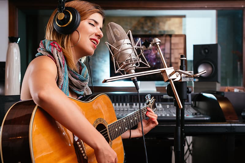 Woman playing a guitar and singing into a microphone ina recording studio.