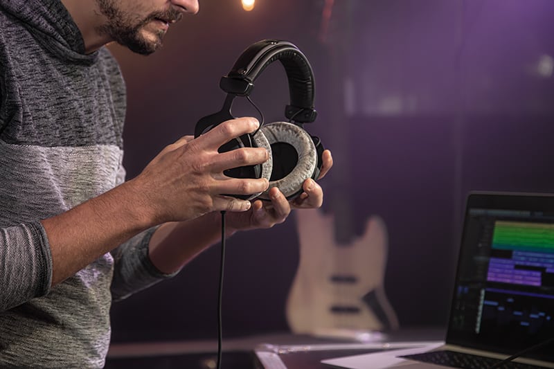 Man holding headphones with a laptop and guitar in the background.