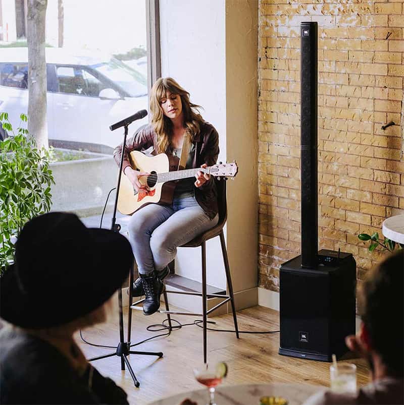 A female singer playing a guitar and using a JBL PRX ONE in a small cafe.