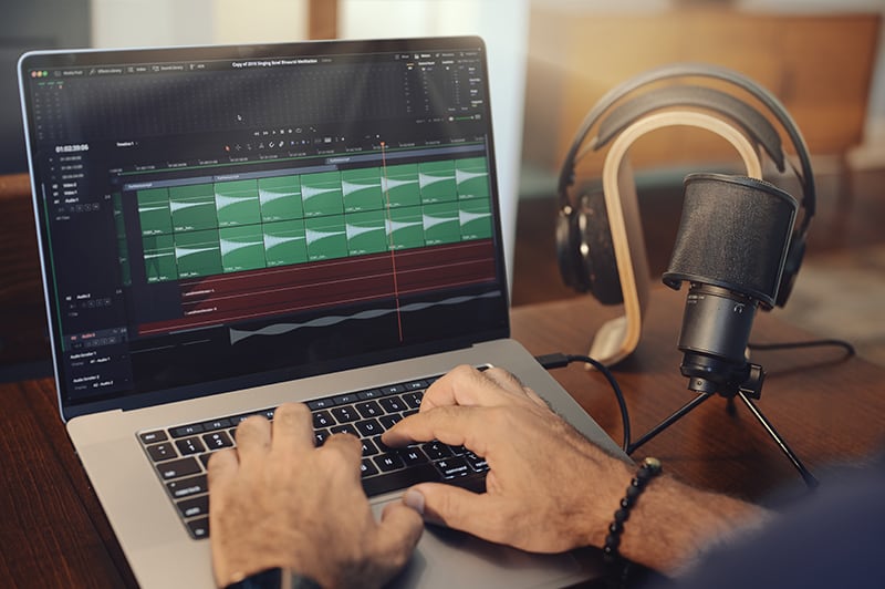 Editing audio on laptop computer with hands on the keyboard. Microphone and headphones on the right side of the laptop.