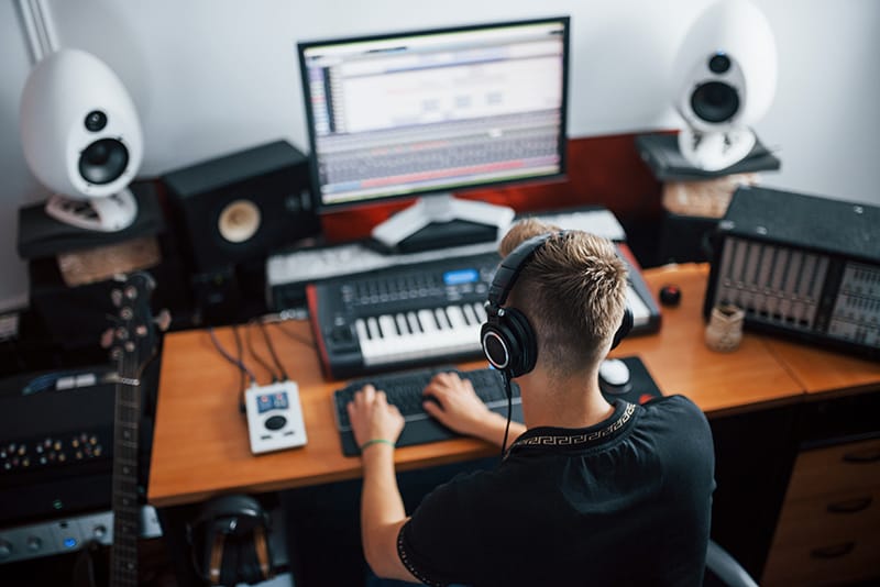 Music producer with headphones on sitting at his desk with a keyboard, guitar and speakers.