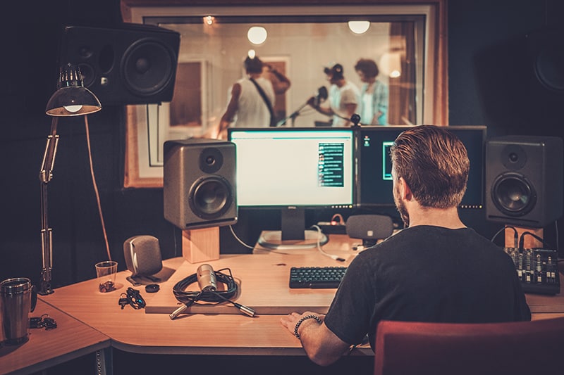 Man sitting at a desk in a professional music recording studio with people in the sound booth.