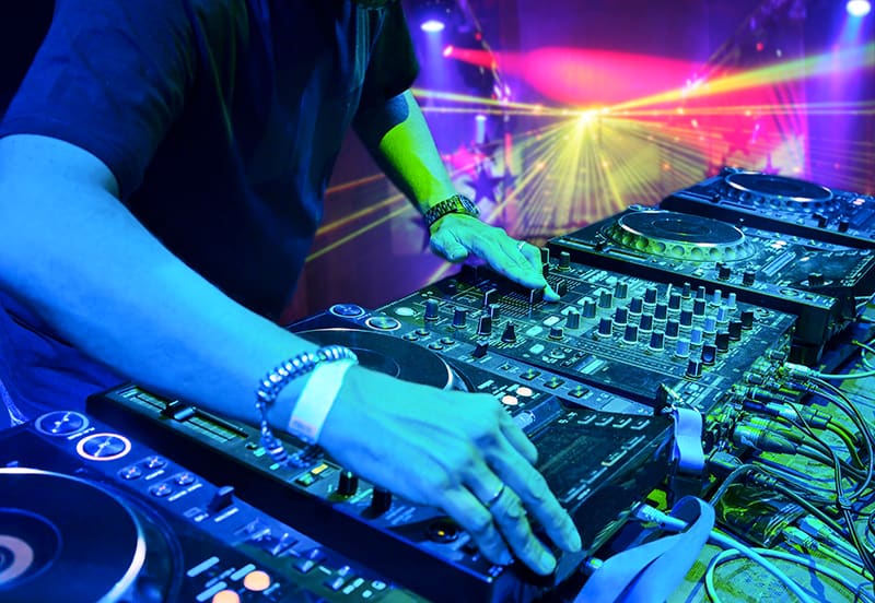 DJ using a DJ mixer and media players in a club with lights in the background.