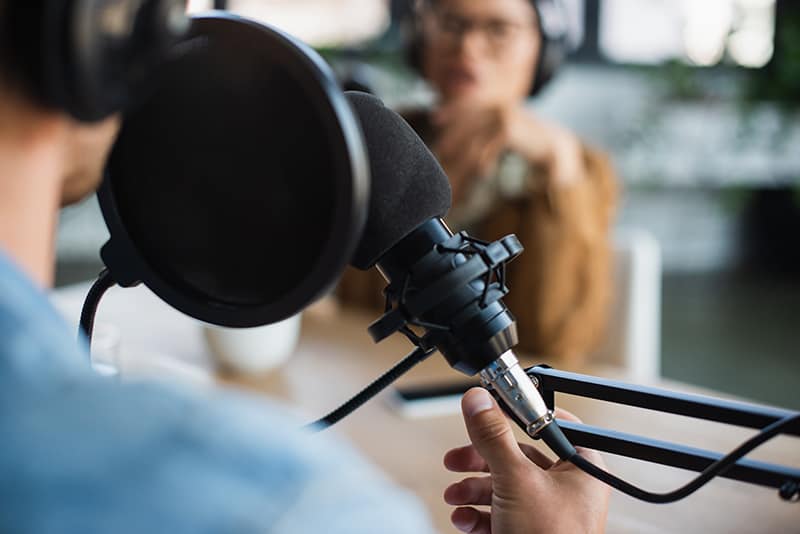 Man wearing headphones in front of a microphone with a blurred person in the background.